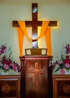 A wooden pulpit with a cross design is adorned with a bright yellow cloth draped over a larger wooden cross. Flower arrangements with pink and white blossoms are placed on each side, contributing to a serene and solemn atmosphere.