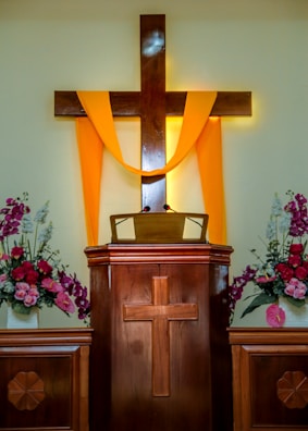 A wooden pulpit with a cross design is adorned with a bright yellow cloth draped over a larger wooden cross. Flower arrangements with pink and white blossoms are placed on each side, contributing to a serene and solemn atmosphere.