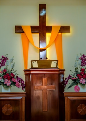A wooden pulpit with a cross design is adorned with a bright yellow cloth draped over a larger wooden cross. Flower arrangements with pink and white blossoms are placed on each side, contributing to a serene and solemn atmosphere.