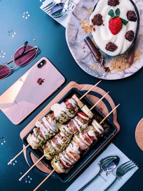 Various desserts are beautifully arranged on a table. A dessert on skewers coated with chocolate and white sauce sprinkled with pistachios is presented on a wooden tray. Beside it, a dessert with a cream base is topped with a fresh strawberry and chocolate pieces, accompanied by chocolate-filled syringes. Nearby are sunglasses, a pink phone, and utensils.