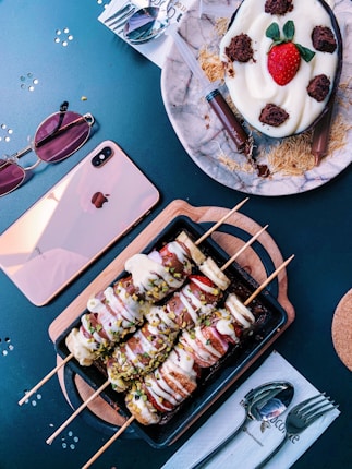 Various desserts are beautifully arranged on a table. A dessert on skewers coated with chocolate and white sauce sprinkled with pistachios is presented on a wooden tray. Beside it, a dessert with a cream base is topped with a fresh strawberry and chocolate pieces, accompanied by chocolate-filled syringes. Nearby are sunglasses, a pink phone, and utensils.