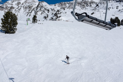 A skier glides down a snowy mountain slope beneath a clear blue sky. Pine trees are scattered throughout the landscape, providing a backdrop against the steep, snow-covered peaks. A ski lift chair is seen overhead, slightly off to the side.