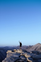 A person stands triumphantly on a rugged mountain peak, raising one arm in a gesture of victory. The expansive landscape behind features a range of distant mountains under a clear blue sky, evoking a sense of freedom and accomplishment.