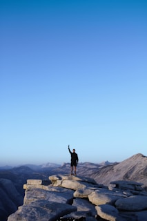 A person stands triumphantly on a rugged mountain peak, raising one arm in a gesture of victory. The expansive landscape behind features a range of distant mountains under a clear blue sky, evoking a sense of freedom and accomplishment.