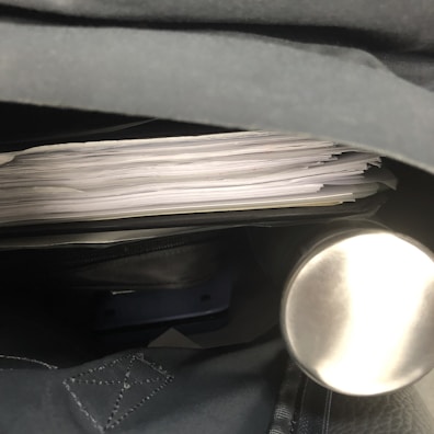 A stack of papers is stored inside a dark-colored bag, featuring a metal cylindrical object, likely a thermos, on one side.
