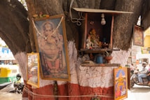 A large tree trunk serves as a makeshift shrine with images of a deity, small statues, and offerings like flowers and bowls. The tree is partially painted with white and red colors, and the shrine includes a light bulb. Surrounding the shrine are various framed religious images, and in the background, a busy street scene with vehicles and people is visible.