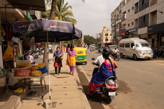 A busy street scene featuring vendors with flowers displayed under Pepsi-branded umbrellas, people walking on the sidewalk, and various vehicles on the road including an auto-rickshaw and a motorcycle with two riders. The backdrop includes several buildings and palm trees.