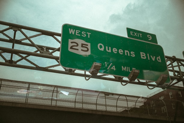 A green highway sign indicating directions towards 'Queens Blvd' with a West 25 route number and Exit 9, seen against a backdrop of a cloudy sky and urban infrastructure.