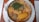 A metal bowl filled with a colorful assortment of food, placed on a white ceramic plate. The dish includes pieces of yellow boiled plantains, meat, a tomato-based sauce, and a topping of leafy green vegetables. A fork is placed beside the plate on a wooden table.