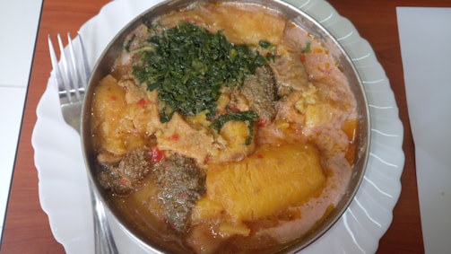 A metal bowl filled with a colorful assortment of food, placed on a white ceramic plate. The dish includes pieces of yellow boiled plantains, meat, a tomato-based sauce, and a topping of leafy green vegetables. A fork is placed beside the plate on a wooden table.
