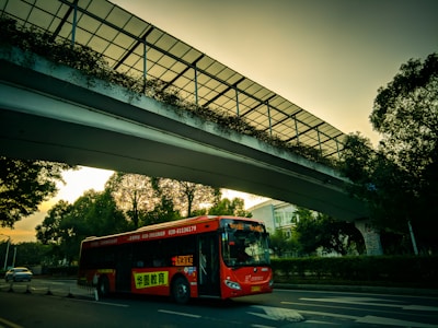 A red bus is traveling on a city road under a modern pedestrian overpass adorned with greenery. The sky exhibits an early evening hue, casting a warm glow overall. Trees line the road, and a car is visible in the background.