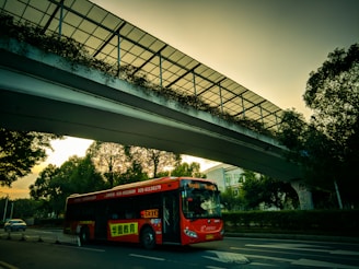A red bus is traveling on a city road under a modern pedestrian overpass adorned with greenery. The sky exhibits an early evening hue, casting a warm glow overall. Trees line the road, and a car is visible in the background.