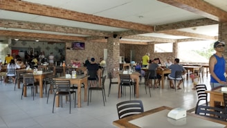 A spacious restaurant area with several tables and chairs, occupied by people dining or socializing. The space has a modern look with exposed brick walls and tiled flooring. Some individuals are watching a television mounted on the wall.