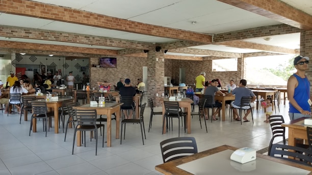 A spacious restaurant area with several tables and chairs, occupied by people dining or socializing. The space has a modern look with exposed brick walls and tiled flooring. Some individuals are watching a television mounted on the wall.