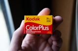 A hand holds a box of Kodak ColorPlus film, displaying the brand name prominently in red and yellow colors. The packaging features details such as 36 exposures and ISO 200. The background is softly blurred, drawing focus to the film box.