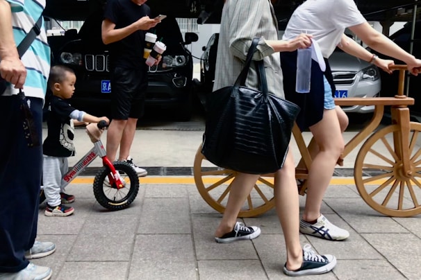 A young child is holding a small balance bike with one hand while standing next to an adult. The adult, dressed in a black outfit, is holding a smartphone and carrying two drinks. Other adults are walking nearby, one with a large black bag, and another holding a large wooden bicycle. Vehicles are parked in the background, and the scene takes place on a paved sidewalk.