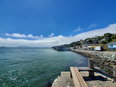 A coastal townscape features a calm blue sea with gentle waves. The skyline of a distant city can be seen across the water, partially shrouded by low-hanging clouds. A rocky seawall runs parallel to a walkway lined with colorful buildings of a quaint town, nestled against a lush green hillside. A wooden bench is situated in the foreground, providing a peaceful spot to enjoy the scenic view.
