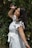 A woman with long dark hair smiles joyfully while reaching towards a pink flower among dense green foliage. She is wearing a white dress with ruffled sleeves and has bracelets on her wrist. The background is filled with leaves and flowers, creating a natural, serene setting.