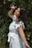 A woman with long dark hair smiles joyfully while reaching towards a pink flower among dense green foliage. She is wearing a white dress with ruffled sleeves and has bracelets on her wrist. The background is filled with leaves and flowers, creating a natural, serene setting.