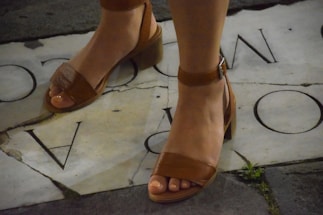A pair of feet wearing brown leather open-toe sandals with ankle straps and block heels, standing on a stone pavement that has partial letters inscribed on it. There is some grass growing between the stones.
