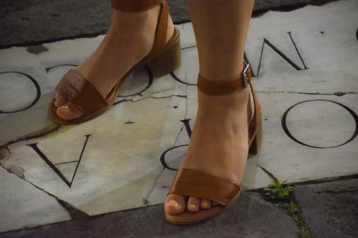 A pair of feet wearing brown leather open-toe sandals with ankle straps and block heels, standing on a stone pavement that has partial letters inscribed on it. There is some grass growing between the stones.