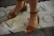 A pair of feet wearing brown leather open-toe sandals with ankle straps and block heels, standing on a stone pavement that has partial letters inscribed on it. There is some grass growing between the stones.