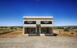 A small, isolated building resembling a boutique store labeled 'PRADA' sits in a barren desert landscape. The building has a minimalist design with large display windows showcasing merchandise. It is surrounded by a dusty, gravel terrain with sparse vegetation and open sky. The setting evokes a sense of surreal juxtaposition between high fashion and the remote, arid environment.
