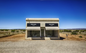 A small, isolated building resembling a boutique store labeled 'PRADA' sits in a barren desert landscape. The building has a minimalist design with large display windows showcasing merchandise. It is surrounded by a dusty, gravel terrain with sparse vegetation and open sky. The setting evokes a sense of surreal juxtaposition between high fashion and the remote, arid environment.
