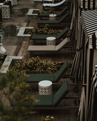 Several lounge chairs with dark green cushions are arranged neatly on a wooden deck surrounded by small planters with lush foliage. Each chair is paired with a small, decorative white side table. Canopies with black and white stripes line one side, adding style to the setting.