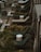 Several lounge chairs with dark green cushions are arranged neatly on a wooden deck surrounded by small planters with lush foliage. Each chair is paired with a small, decorative white side table. Canopies with black and white stripes line one side, adding style to the setting.