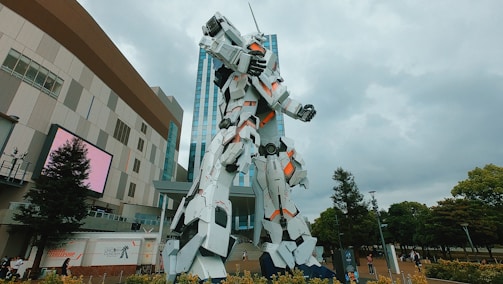 A large, white, humanoid robot statue stands prominently in front of a modern building. The robot features orange accent lighting and mechanical details. Surrounding the area are trees and people walking, suggesting an urban environment.