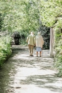 An elderly couple is walking down a sunlit path surrounded by lush greenery. The trees and bushes form a canopy overhead, creating a serene and peaceful atmosphere. The couple walks side by side, with one using a cane for support.