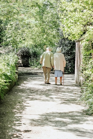 An elderly couple is walking down a sunlit path surrounded by lush greenery. The trees and bushes form a canopy overhead, creating a serene and peaceful atmosphere. The couple walks side by side, with one using a cane for support.