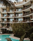 A modern apartment building with large glass windows and balconies adorned with potted plants. The structure features curved architectural elements. In the foreground, a pool surrounded by lush greenery and palm leaves adds a tropical atmosphere.