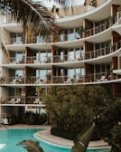 A modern apartment building with large glass windows and balconies adorned with potted plants. The structure features curved architectural elements. In the foreground, a pool surrounded by lush greenery and palm leaves adds a tropical atmosphere.