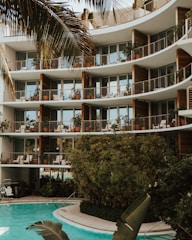 A modern apartment building with large glass windows and balconies adorned with potted plants. The structure features curved architectural elements. In the foreground, a pool surrounded by lush greenery and palm leaves adds a tropical atmosphere.