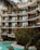 A modern apartment building with large glass windows and balconies adorned with potted plants. The structure features curved architectural elements. In the foreground, a pool surrounded by lush greenery and palm leaves adds a tropical atmosphere.