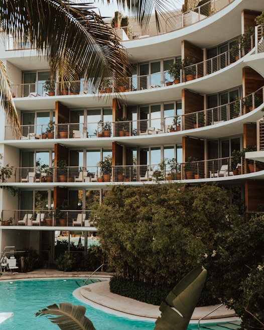 A modern apartment building with large glass windows and balconies adorned with potted plants. The structure features curved architectural elements. In the foreground, a pool surrounded by lush greenery and palm leaves adds a tropical atmosphere.