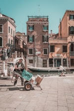A sunlit street scene featuring an individual pushing a hand truck loaded with various boxes and a teal plastic container. The background showcases traditional Venetian architecture, complete with colorful facades and shuttered windows. A canal runs alongside the street, with a small boat tied to the bank. Nearby, people are casually standing and sitting, enjoying the day.