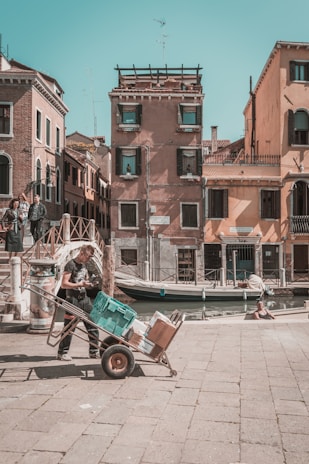 A sunlit street scene featuring an individual pushing a hand truck loaded with various boxes and a teal plastic container. The background showcases traditional Venetian architecture, complete with colorful facades and shuttered windows. A canal runs alongside the street, with a small boat tied to the bank. Nearby, people are casually standing and sitting, enjoying the day.