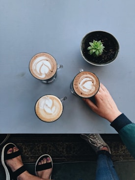 Three cups of coffee with latte art sit on a table next to a small potted succulent. A hand is reaching for one of the cups, and two pairs of feet are visible, one with sandals and painted toenails and the other with sneakers.