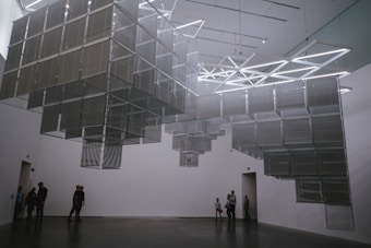 A modern art installation featuring a series of geometric metal structures suspended from the ceiling in a gallery with a white interior. Several people stand and walk around, observing the installation. The lighting casts soft shadows on the walls and floor, creating a subtle interplay between light and architecture.