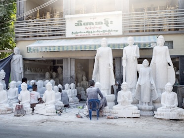 Several artisans are working on large white statues of religious figures, which are arranged outside a workshop. The workshop has a sign in a foreign language and there are more statues placed inside the building. The scene includes tools and equipment scattered on the ground, and some of the artisans are kneeling or sitting on small stools as they work on the sculptures.