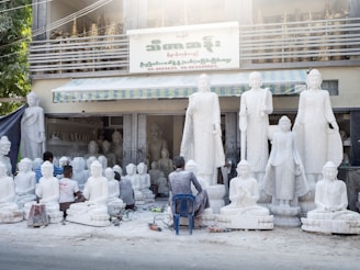 Several artisans are working on large white statues of religious figures, which are arranged outside a workshop. The workshop has a sign in a foreign language and there are more statues placed inside the building. The scene includes tools and equipment scattered on the ground, and some of the artisans are kneeling or sitting on small stools as they work on the sculptures.
