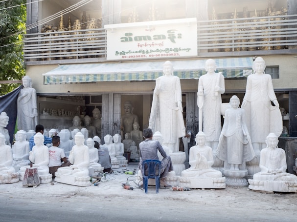 Several artisans are working on large white statues of religious figures, which are arranged outside a workshop. The workshop has a sign in a foreign language and there are more statues placed inside the building. The scene includes tools and equipment scattered on the ground, and some of the artisans are kneeling or sitting on small stools as they work on the sculptures.