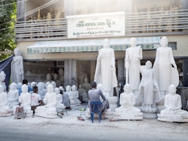 Several artisans are working on large white statues of religious figures, which are arranged outside a workshop. The workshop has a sign in a foreign language and there are more statues placed inside the building. The scene includes tools and equipment scattered on the ground, and some of the artisans are kneeling or sitting on small stools as they work on the sculptures.