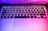 Close-up of a keyboard with purple and pink backlighting reflecting on a matte black desk.
