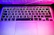 Close-up of a keyboard with purple and pink backlighting reflecting on a matte black desk.