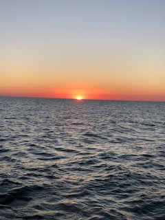 A serene ocean scene with gentle waves under a clear sky during a sunset. Vibrant hues of orange and pink fill the horizon as the sun dips below the waterline, creating a peaceful and calm atmosphere.