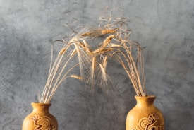 Two ornately decorated ceramic vases are positioned side by side against a textured gray background. Each vase holds golden stalks of wheat that curve gracefully, creating an arch above the vases. The pottery features artistic designs etched into its surface, adding to the rustic and natural aesthetic.
